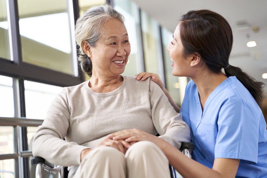 An elderly senior living female patient sitting in a wheelchair as a female nursing assistant kneels beside her