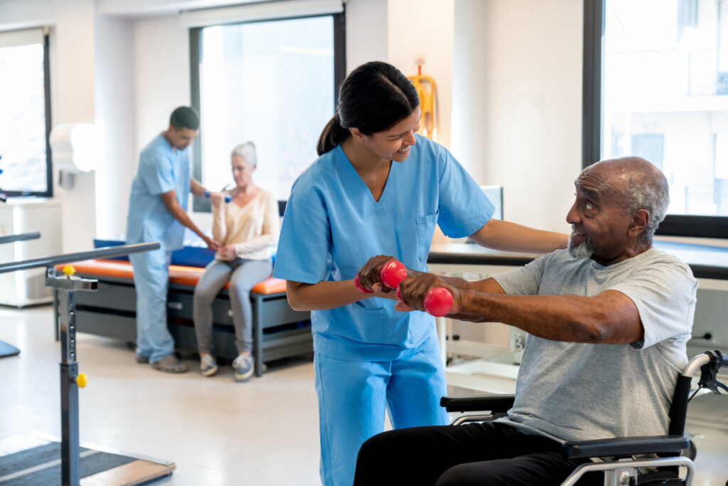 A senior man sitting in a wheelchair holding small red weights as a female nursing assistant helps him